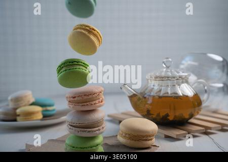 pastel macarons levitating and stacked beside a glass teapot of amber tea on a wooden trivet, soft natural light on a minimalist tabletop studio scene Stock Photo