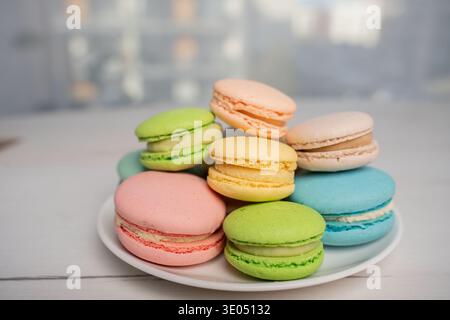 close-up of pastel-colored macarons stacked on a white plate on a pale wooden tabletop in a soft-focus indoor studio kitchen, featuring delicate frenc Stock Photo