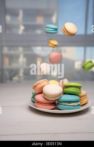 colorful pastel macarons stacked on a white plate beside a sunlit window in a minimal kitchen or apartment setting, multicolored french confections ca Stock Photo