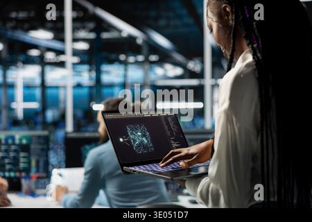 Technician in data center using machine learning LLM assistant to do maintenance on equipment rigs. African american woman in server hub using large language model chatbot to do backups Stock Photo