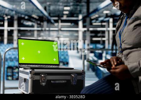 Mockup laptop next to female engineer in server hub troubleshooting hardware and software issues. African american data center worker using green screen notebook, optimizing system performance Stock Photo