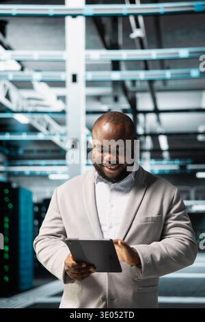 Cheerful data center technician using tablet to protect servers from exploitable software vulnerabilities, auditing systems. Jolly cybersecurity professional looking for bugs slowing down servers Stock Photo