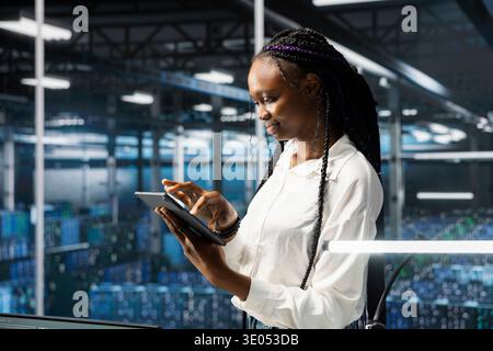 Smiling IT professional using tablet in server farm with hardware supporting data mining. Happy woman in data center using device to do maintenance on racks storing blockchain records Stock Photo