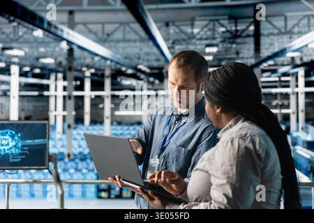 Teamworking engineers in data center deploying infrastructure for efficient AI data analysis. Coworkers using laptop in server farm, managing storage systems for AI training datasets Stock Photo