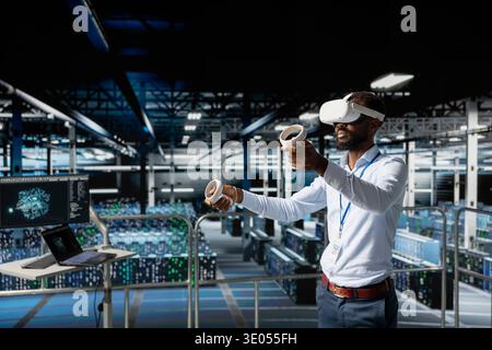 Software developer managing performance outputs with virtual reality tech on elevated industrial platform in data center, handling server management using VR glasses. Supercomputers. Stock Photo