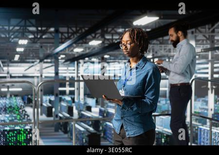 Woman engineer on data center industrial platform performing system maintenance, high tech. IT professional using laptop to do upgrades, resolving bugs and enhancing infrastructure stability. Stock Photo