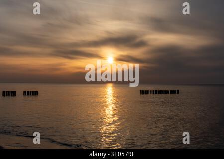 Dramatic golden sunset over calm Baltic sea with silhouette of old wooden groins reflected in glowing water.  Stock Photo