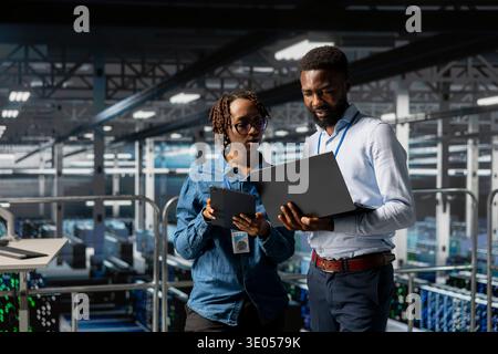 Black software developers on industrial platform analyzing server rigs algorithms in data center, using laptop and tablet. Server room IT specialists collaborating on finetuning PC configurations. Stock Photo