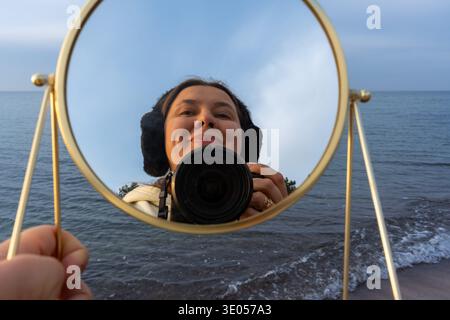 Smiling woman photographer taking selfie in round golden mirror held against windy sea on overcast day.  Stock Photo