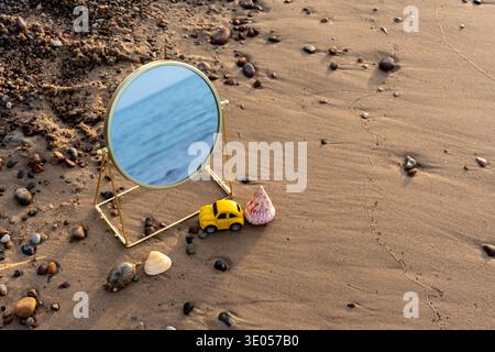 Round golden mirror with sea reflection standing on sandy beach beside yellow toy car and seashells with pebbles.  Stock Photo