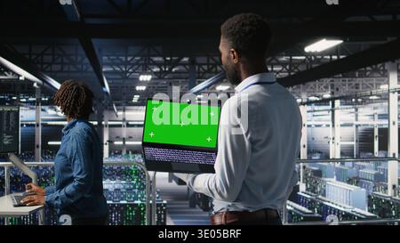 Technicians on data center industrial platform analyzing performance outputs on green screen laptop. Server hub engineering team finetuning server rig configurations using mockup notebook, Stock Photo
