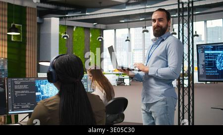 Jolly programmer coding AI algorithms on laptop to debug software issues and improving system stability. Smiling employee in tech startup office configuring settings using notebook, camera A Stock Photo