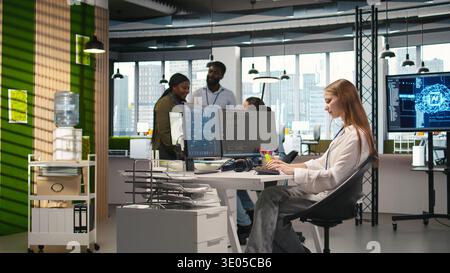 Manager supervising young trainee in office using AI technology, monitoring performance dashboards. Team leader overseeing work done by junior dev tuning neural networks using tools on PC, camera A Stock Photo