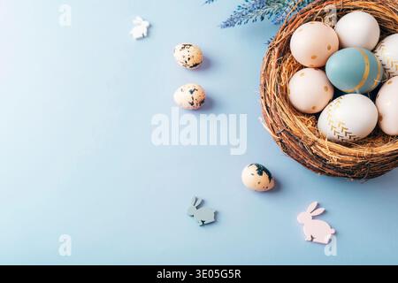 Easter eggs in nest with rabbit decorations on blue background. Top view, flat lay, copy space. Stock Photo
