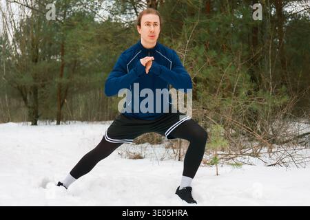 Fit young man performing a side lunge stretch in a snowy winter forest. He wears athletic gear for outdoor cold weather training and fitness. High qua Stock Photo
