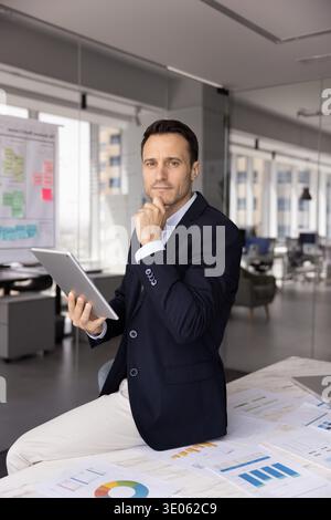 Pensive businessman sitting on desk looking at camera holding tablet Stock Photo