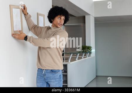 Glancing right, man in beige top aligning two frames to match mounted piece on white wall. Minimalist, contemporary, neutral, interior, apartment, dec Stock Photo