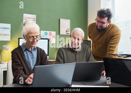 Senior Caucasian woman and senior Caucasian man participating in computer literacy course, using laptops while young adult Caucasian man instructor assisting and explaining technology concepts Stock Photo