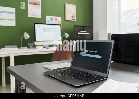 Modern classroom featuring open laptop with web browser on screen positioned on desk in foreground, desktop computers and educational posters visible in background Stock Photo