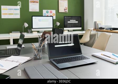 Modern classroom featuring open laptop displaying web browser on desk, multiple desktop computers in background, educational materials and stationery suggesting computer literacy course setting Stock Photo
