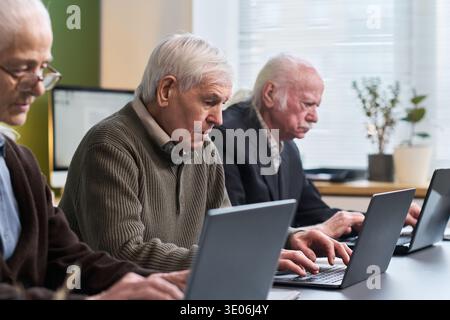 Group of senior Caucasian men sitting in classroom setting using laptop computers, participating in computer literacy course, focusing on learning digital skills together Stock Photo