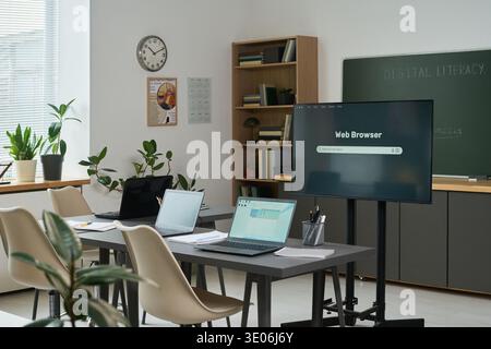 Empty classroom with several laptops and large monitor displaying web browser on table, digital literacy course materials visible on chalkboard Stock Photo