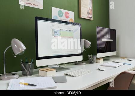 Modern classroom featuring two desktop computers on desks with notebooks, pens, and lamps arranged for computer literacy course training, educational posters on green wall Stock Photo