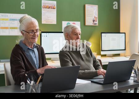 Senior Caucasian woman and senior Caucasian man sitting at desks using laptops during computer literacy course, both focused on screens with desktop computers visible in background Stock Photo