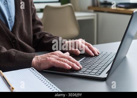 Unknown elderly woman typing on laptop during computer literacy course, hands visible on keyboard, spiral notebook and pencil on desk, participating in digital skills training Stock Photo