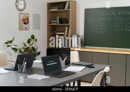 Empty classroom with several laptops on table displaying web browser error, notebooks and pens scattered around, chalkboard in background showing digital literacy course instructions Stock Photo