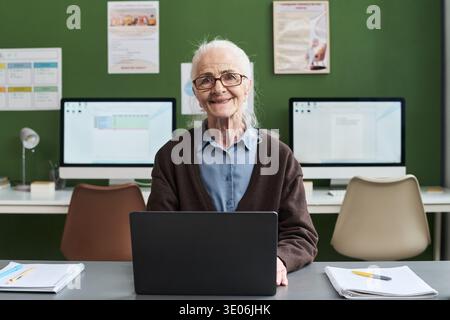 Portrait of senior Caucasian woman smiling, while sitting at desk using laptop during computer literacy course with desktop monitors and notebooks visible in classroom setting Stock Photo