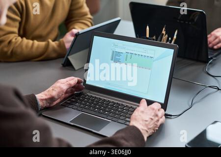 Senior woman using laptop during computer literacy course, hands typing on keyboard while following spreadsheet tutorial, other participants working on tablets in background Stock Photo