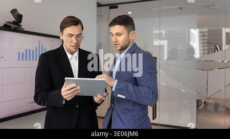 Two businessmen standing in office reviewing information on digital tablet Stock Photo