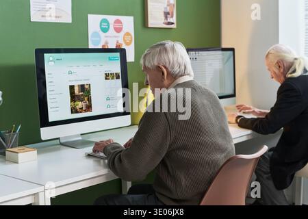 Senior Caucasian man using desktop computer during computer literacy course, sitting at desk and typing on keyboard, another senior Caucasian man working on laptop in background Stock Photo