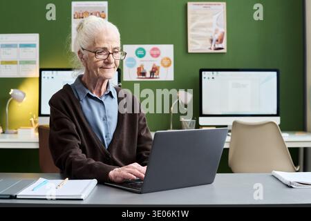 Senior Caucasian woman sitting at desk using laptop computer during computer literacy course, typing on keyboard with focused expression, classroom with desktop monitors in background Stock Photo