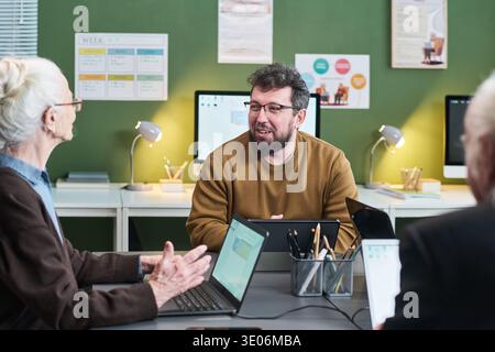 Middle aged Caucasian man teaching senior Caucasian woman and senior Caucasian man using laptops and tablet in computer literacy course classroom, with digital devices visible Stock Photo