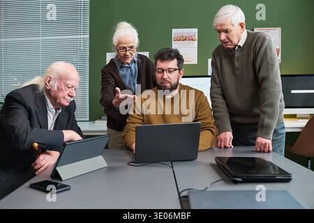 Group of senior Caucasian men and woman participating in computer literacy course, sitting around table using laptops and tablet, woman gesturing while discussing technology skills Stock Photo