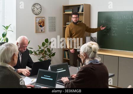 Middle aged Caucasian man teaching senior Caucasian men and women computer literacy course, standing by chalkboard with digital literacy instructions while seniors using laptops Stock Photo