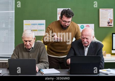 Middle aged Caucasian man instructing two senior Caucasian men using laptops during computer literacy course, seniors focusing on screens while teacher explaining concept Stock Photo