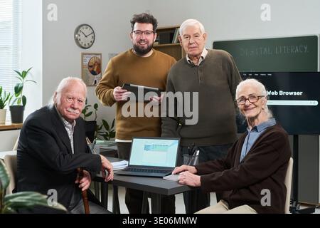 Group of middle aged Caucasian man, senior Caucasian man, senior Caucasian woman and young adult Caucasian man participating in computer literacy course using laptop and tablet in classroom Stock Photo