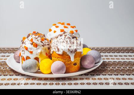 A festive arrangement of elaborately decorated Easter kulich cakes and dyed eggs presented on a white plate atop a patterned tablecloth Stock Photo
