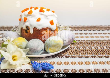 A beautifully decorated Easter cake with white icing and dried fruit sits beside intricately painted eggs and spring blossoms on a patterned cloth Stock Photo