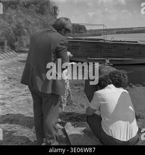 An agricultural manager wearing a formal suit carefully writes in a small notebook while observing dedicated workers closely examining the fresh aquatic catch on the muddy pond bank at the large Donrybkombinat facility in the Sloviansk district during the 1980s. This engaging archival image beautifully captures the strict administrative control, essential daily accounting procedures, and authentic manual field labor typical of massive Soviet collective farms, representing a truly genuine record of a peaceful Donbass before the war. Stock Photo