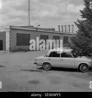 A classic archival black and white photograph from the 1980s shows a typical Soviet passenger car parked next to a large industrial facility at the Donrybkombinat agricultural enterprise in the Slavyansk district. The utilitarian scene features a long brick building with characteristic glass block windows, tall ventilation chimneys on the roof, and lush green trees near the paved courtyard. This authentic snapshot perfectly documents the functional rural infrastructure, transportation, and everyday working life in the peaceful Donbass before the war. Stock Photo