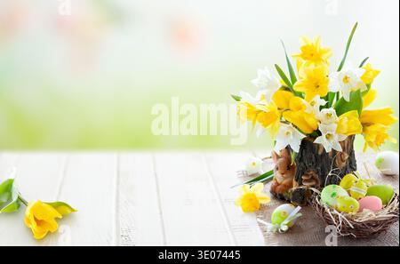 A vase of yellow and white flowers sits on a wooden table. The flowers are surrounded by a basket of eggs and a bird's nest. Concept of warmth and com Stock Photo