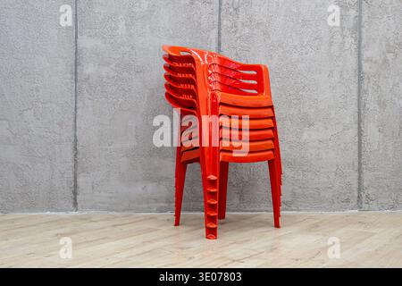 Three-quarter view of glossy orange plastic stacking chairs organized vertically against gray concrete wall on wooden floor for efficient storage, sel Stock Photo