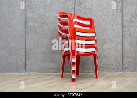 Three-quarter view of glossy orange and white plastic stacking chairs alternating in vertical stack against gray concrete wall on wooden floor, select Stock Photo