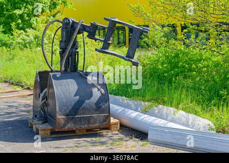 Black excavator bucket attachment with hydraulic arm placed on a wooden pallet near green bushes and construction materials outdoors. Stock Photo