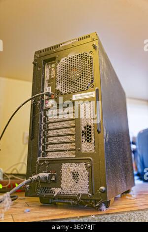 A dusty computer tower with clogged ventilation holes and cables connected, sitting on a wooden floor in an indoor setting with visible dust accumulat Stock Photo
