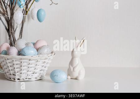 Easter tree with pastel speckled eggs hanging on spring branches in glass vase. White ceramic bunny and basket with painted eggs on white background. Stock Photo